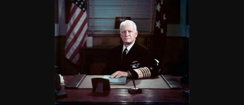 Fleet Admiral Chester W. Nimitz seated at his desk as Chief of Naval Operations, flanked by American flags